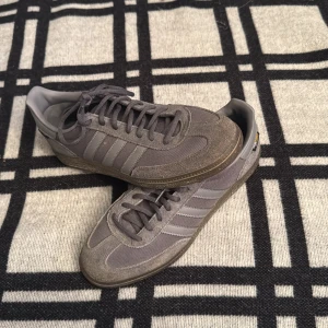 Grå adidas Samba sneakers - Säljer ett par grå adidas Samba sneakers med klassiska tre ränder på sidan. Skorna har ovandel i mocka och textil, snörning och platt sula. Perfekta för dig som gillar retrostil och vill ha något som sticker ut men ändå är stilrent.