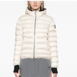 Vit dunjacka från Colmar - Säljer en fräsch vit dunjacka från Colmar med huva och svart logga på ärmen. Jackan har puffad design, dragkedja framtill och långa ärmar med svarta muddar. Perfekt för kalla dagar och har en clean, modern look.