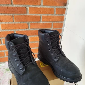 Svarta Timberland boots i skinn - Säljer ett par svarta Timberland boots med robust snörning och grov platt sula. Skorna är tillverkade i äkta skinn med vadderad krage för extra komfort. Perfekta för höst och vinter, med klassisk rund tå och diskret Timberland-logga på sidan.