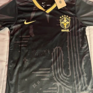 Svart Brasilien fotbollströja Nike - Snygg svart Brasilien fotbollströja från Nike med gula detaljer och tryckt CBF-märke på bröstet. Tröjan har korta ärmar, V-ringad krage med gul kant och är tillverkad i Dri-FIT-material som andas. Unik design med subtila grafiska mönster på framsidan.