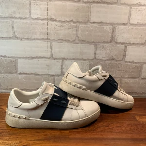 Valentino Garavani Open sneakers vit/blå - Snygga Valentino Garavani Open sneakers i vitt skinn med bred marinblå läderdetalj över snörningen. Klassisk rund tå, platt sula och ikoniska nitar längs hälen. Perfekta för dig som vill ha en stilren men ändå lyxig look.