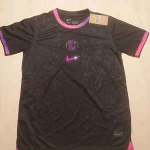 PSG x Nike svart fotbollströja - Svart Paris Saint-Germain fotbollströja från Nike med lila och rosa detaljer på krage och ärmslut. Tröjan har ett diskret blommönster i tyget, PSG-logga och en rosa Nike swoosh framtill. Tillverkad i lätt och ventilerande Dri-FIT-material.