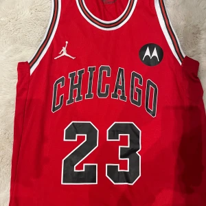 Chicago Bulls Jordan 23 basketlinne Nike - Röd Chicago Bulls baskettröja från Nike med Jordan-nummer 23, klassisk svart och vit kant runt hals och ärmhål. Broderad Jumpman-logga och Motorola-märke på bröstet. Tillverkad i ventilerande meshmaterial, perfekt för basket eller streetwear.