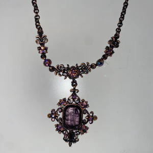 Vintage halsband med lila sten - Unikt vintage halsband med antik bronsfärgad kedja och detaljerad medaljong. Medaljongen har en stor lila sten i mitten, omgiven av glittriga små stenar i lila och guld. Kedjan har dekorativa blommönster och gnistrande accenter som ger ett lyxigt och romantiskt uttryck.