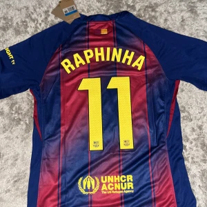 FC Barcelona Raphinha matchtröja Nike M - Säljer en officiell FC Barcelona matchtröja med Raphinha 11 på ryggen. Tröjan är kortärmad, tillverkad i Dri-FIT material och har klassiska blå och röda ränder, klubbmärke, Nike-logga och sponsorer i gult. Perfekt för dig som älskar Barca och vill sticka ut på läktaren.