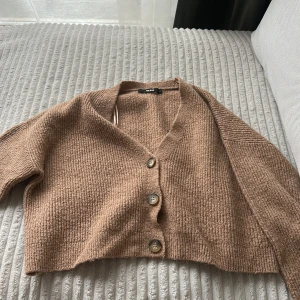 Beige stickad kofta  - Mysig beige stickad kofta med tre knappar framtill. Koftan är croppad och har en avslappnad passform med v-ringning. Perfekt att slänga över en t-shirt eller linne för en chill look.