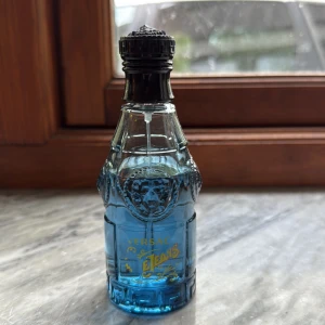 Versace Blue Jeans parfymflaska - Stilren parfym från Versace med blått innehåll och ikonisk design. Perfekt för dig som vill sticka ut med en klassisk och ungdomlig touch. Flaskans detaljer ger en exklusiv känsla.