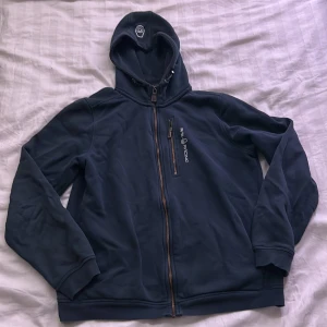 Marinblå hoodie från Sail Racing - Marinblå hoodie från Sail Racing med dragkedja framtill och bröstficka med dragkedja. Broderad logga på bröstet och huva med tryckt detalj. Mjuk insida och ribbade muddar vid ärmslut och nederkant. Perfekt för kyliga dagar.