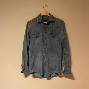 Slim fit jeansskjorta från Dressmann - Säljer en snygg ljusblå jeansskjorta från Dressmann i slim fit-modell. Skjortan har två bröstfickor med knappar och klassisk krage. Perfekt för dig som gillar en stilren och avslappnad look.