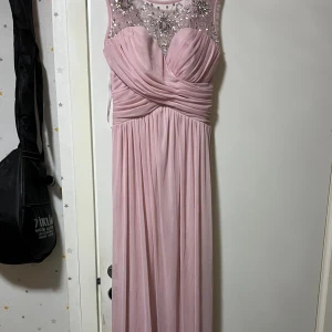Ljusrosa långklänning med pärldetaljer - Ljusrosa långklänning med vackert draperad överdel och transparent mesh upptill, dekorerad med glittrande pärlor och stenar. Klänningen har markerad midja, lätt utställd kjol och är ärmlös. Perfekt för bal eller speciella tillfällen. Men de råkar spillas lite sås på den vid bröstet, priset går att diskutera 