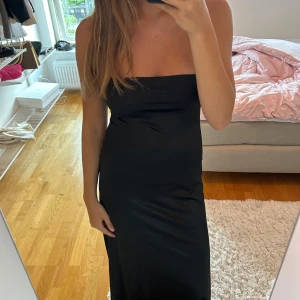 Svart axelbandslös långklänning - Säljer en stilren svart långklänning med axelbandslös design. Klänningen har en rak och figurnära passform som ger en snygg siluett. Materialet är mjukt och har en lätt glansig yta som känns lyxig. Perfekt för dig som gillar minimalistisk och elegant stil.