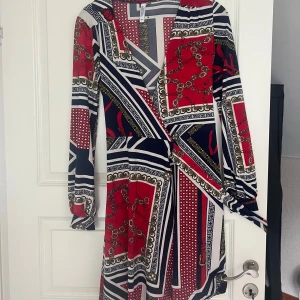 Mönstrad omlottklänning från bubbleroom - Säljer en färgstark omlottklänning med lång ärm och coolt mönster i rött, marinblått, vitt och guld. Klänningen har v-ringning och knyt i midjan, perfekt för dig som gillar statement-plagg och snygga detaljer.