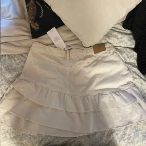 Vit volangkjol från Gina Tricot - Supersöt vit kortkjol från Gina Tricot med volangdetaljer och klassisk jeanslook. Kjolen har fickor fram och bak, bälteshällor och stängs med dragkedja och knapp. Säljer för att den är för stor för mig.