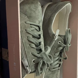 sneakers från Legero - Snygga sneakers från Legero i grå mocka med vita sulor och svarta detaljer undertill. Skorna har snörning framtill och dragkedja på sidan för extra stil och smidighet. Perfekta för dig som gillar en clean och modern look.