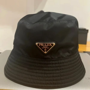 Svart bucket hat från Prada - Stilren svart bucket hat från Prada med ikonisk triangelformad logotyp framtill i metall och läderdetaljer. Hatten är tillverkad i glansig syntet och har klassisk form med rundad kulle och nedvikt brätte. Perfekt accessoar för en trendig look.