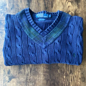 Polo Ralph Lauren Pullover - Polo Ralph Lauren kabelstickad tröja i mörkblått med gröna detaljer vid kragen. Storlek Small, 100% bomull. Klassisk V-ringad modell med kabelstickning och diskret broderad logga. Bra skick och perfekt för både vardag och mer uppklädda tillfällen. 