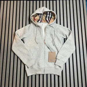 Grå hoodie från Burberry med dragkedja - Snygg grå hoodie från Burberry med klassiskt rutigt foder i huvan. Tröjan har dragkedja framtill, snörning i huvan och ribbade muddar. Perfekt för dig som vill ha en stilren och exklusiv look med en touch av Burberrys ikoniska mönster.