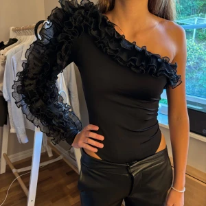 Svart oneshoulder body med volang - Säljer en svart oneshoulder body med en lång ärm och stora volangdetaljer längs ärmen. Perfekta fest toppen!!