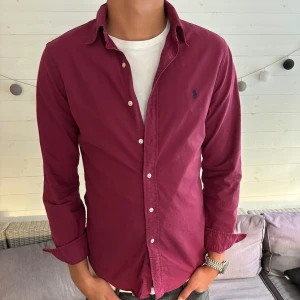 Vinröd skjorta från Ralph Lauren - Säljer en vinröd skjorta från Ralph Lauren med klassisk krage och knappar framtill. Skjortan har lång ärm och den ikoniska blå loggan broderad på bröstet. Tillverkad i mjuk bomull och har en normal passform. Perfekt för dig som vill ha en stilren look.