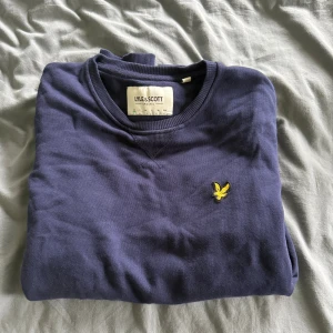 Blå sweatshirt från Lyle & Scott - En stilren blå sweatshirt från Lyle & Scott med klassisk rund hals och deras ikoniska gula fågel-logga broderad på bröstet. Tröjan är i mjuk bomull och har ribbade muddar vid ärmslut och nederkant. Perfekt för en chill och avslappnad look.