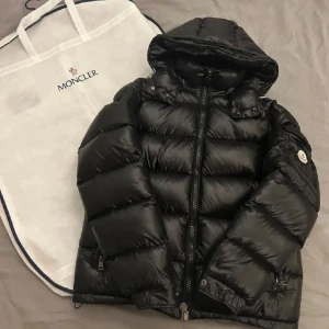 Svart Moncler dunjacka med huva - Snygg svart dunjacka från Moncler med glansig finish och klassisk pufferstil. Jackan har huva, dragkedja framtill, två fickor med dragkedja och Moncler-logga på ärmen. Insidan har ett unikt serietidningsmärke. Perfekt för kalla dagar när du vill hålla stilen.