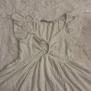 Vit kortklänning med volangärmar XS - Superfin vit kortklänning i luftig bomull med volangärmar och knytning framtill. Klänningen har en söt cut-out detalj vid bysten och ett lätt rynkat tyg som ger en avslappnad och somrig vibe. Perfekt för varma dagar och picknickar.