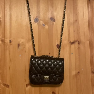 Svart quiltad axelväska med guldiga detaljer - Snygg svart axelväska i quiltat skinnimitation med guldfärgad kedjerem och vridlås framtill. Väskan har en klassisk, stilren look och passar perfekt när du vill lyfta din outfit med en trendig accessoar.