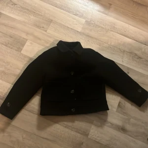 Svart kavajjacka från 157, stl 140 - Säljer en svart kavajjacka från 157 i storlek 140. Jackan har klassisk krage, knäppning med stora knappar framtill och två fickor. Fodrad insida för extra komfort. Perfekt för dig som vill ha en stilren look.