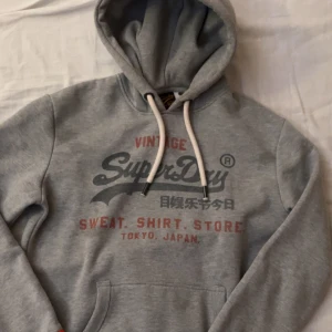 Superdry hoodie  - Säljer en grå hoodie från Superdry med stort tryck på bröstet i svart och rött. Tröjan har huva med vita snören och en klassisk känguruficka framtill. Perfekt för dig som gillar streetwear och vill ha en skön och snygg look.