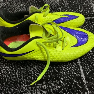Neongula Nike fotbollsskor med lila detaljer - Säljer ett par riktigt coola fotbollsskor från Nike i neongult med lila swoosh och svart dödskalle-tryck på hälen. Skorna har snörning, mönstrad ovansida och dobbar för gräsplan. Insidan är svart och röd, och materialet är syntet. Perfekta för dig som vill sticka ut på planen!