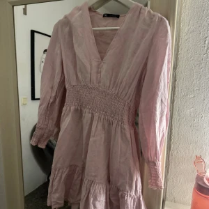 Ljusrosa kortklänning från Zara - Supersöt ljusrosa klänning från Zara med v-ringning, långa ärmar och smock i midjan och vid ärmsluten. Klänningen har volangdetaljer och är tillverkad i ett lätt och luftigt material som passar perfekt till varmare dagar.