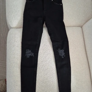 Svarta skinny jeans med slitningar - Svarta skinny jeans med hög midja och slitna detaljer på både knän och bakfickor. Jeansen har klassisk femficksdesign, bälteshällor och dragkedja med knapp. Materialet är stretchigt denim som sitter tight och bekvämt. Helt nya och oanvända jeans utan prislapp.