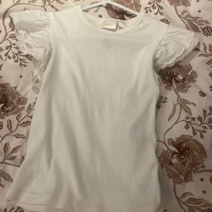 Vit blus med volangärm från Gina Tricot - Supersöt vit blus från Gina Tricot med ribbad kropp och korta volangärmar i broderad bomull. Ärmarna har fina broderade detaljer och vågig kant. Perfekt för dig som gillar en romantisk och fräsch stil. som man ser på första bilden så har den en liten fläck men fläcken syns inte mycket när tröjan är på! därför säljs den för billigare pris💗