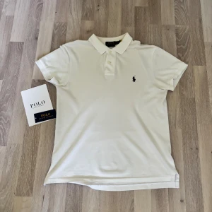 Vit pikétröja Polo Ralph Lauren - Klassisk vit pikétröja från Polo Ralph Lauren i slim fit. Tröjan har korta ärmar, krage med knappar och den ikoniska broderade loggan på bröstet. Tillverkad i bomull för en skön och luftig känsla. Perfekt för en clean och stilren look.