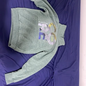 Grön hoodie från Jack & Jones CORE - Säljer en grön hoodie från Jack & Jones CORE med tryck i gult, vitt och blått framtill. Tröjan har hög krage, magficka och långa ärmar. Perfekt för dig som gillar en avslappnad stil och vill ha något bekvämt och snyggt.