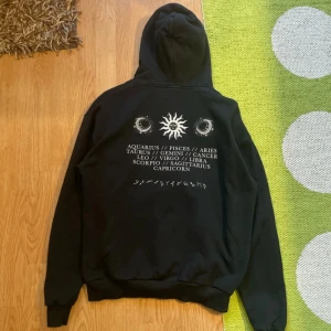 Svart hoodie med stjärntecken från Madlady - Svart hoodie från Madlady med vit tryck av sol, månar och alla stjärntecken på både fram- och baksidan. Tröjan har huva och en stor magficka. Perfekt för dig som gillar astrologi och vill ha en cool och bekväm stil.
