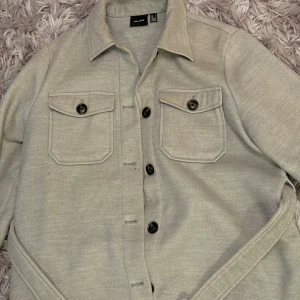 Grå overshirt från Vero Moda S - Stilren overshirt från Vero Moda med klassisk krage, två stora bröstfickor med knappar och snygga detaljer vid midjan. Jackan har långa ärmar och stängs med svarta knappar framtill. Perfekt för lager-på-lager och enkel att matcha.