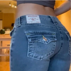 True Religion Becca Bootcut Jeans - Snygga blå bootcut jeans från True Religion, modell Becca Mid Rise Bootcut. Klassisk femficksdesign med ikoniska bakfickor och kontrastsömmar. Jeansen har medelhög midja och är tillverkade i slitstarkt denim med coola detaljer och logopatch bak.