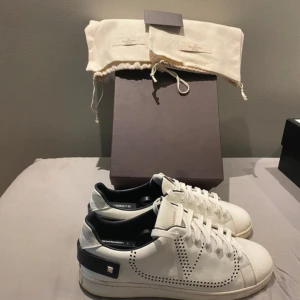 Valentino Garavani vita sneakers 41 - Snygga vita Valentino Garavani sneakers med svarta detaljer och ikoniska nitar på hälen. Skorna har perforerat V-mönster på sidorna, rund tå och platt sula. Tillverkade i skinn och levereras med originalpåse och låda.