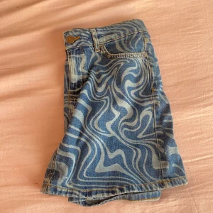 Blå jeanskjol med vågigt mönster Lindex - Säljer en blå jeanskjol från Lindex med unikt vågigt mönster i ljusblått. Kjolen har klassisk femficksdesign, bälteshällor och knapp framtill. Perfekt för dig som vill sticka ut med en cool och trendig denimlook.