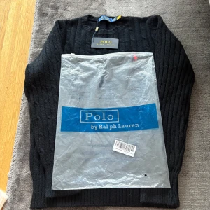 Svart stickad tröja Polo Ralph Lauren - Svart kabelstickad tröja från Polo Ralph Lauren med klassisk röd broderad logga på bröstet. Tröjan har rund halsringning, ribbade muddar och är långärmad. Perfekt för dig som gillar stilren och tidlös design.