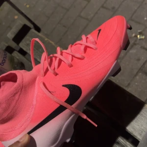 Nike Phantom GT2 rosa fotbollsskor - Nike Phantom GT2 fotbollsskor i en riktigt snygg och iögonfallande rosa färg med svart swoosh. Skorna har snörning, strumpliknande skaft och är tillverkade i syntetmaterial för bästa passform och bollkontroll. Perfekta för dig som vill sticka ut på planen. Köpte dom för 2149kr 