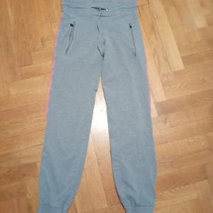 Grå mjukisbyxor från SOC med rosa detalj - Grå mjukisbyxor från SOC med rosa ränder längs sidorna. Byxorna har resår och snörning i midjan, dragkedjeförsedda fickor framtill och muddar vid bensluten. Perfekta för chill eller träning, tillverkade i mjukt bomullsmaterial.