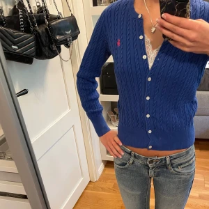 Polo Ralph Lauren kofta - Superfin kabelstickad blå kofta från Ralph Lauren med långa ärmar och knappar framtill. Detaljerad med en rosa broderad logga på bröstet. Står storlek L (12-14) men passar XS. Köpt för 2295kr! 💗