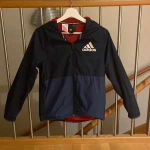 Mörkblå vindjacka från Adidas - Snygg mörkblå vindjacka från Adidas med vit logga på bröstet och röd meshfoder på insidan. Jackan har huva, dragkedja framtill och långärmade ärmar. Perfekt för dig som gillar sportig stil och vill ha något lätt och funktionellt.