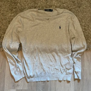 Grå v-ringad tröja Polo Ralph Lauren - Klassisk ljusgrå v-ringad tröja från Polo Ralph Lauren med broderad marinblå logga på bröstet. Tröjan är långärmad och har ribbade muddar vid ärmslut och nederkant. Perfekt för lager-på-lager och enkel att matcha.