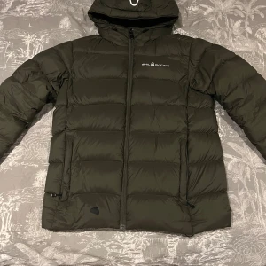 Dunjacka Sail Racing - Säljer en olivgrön dunjacka från Sail Racing med huva och dragkedja framtill. Jackan är vadderad och har två sidofickor med dragkedja. Perfekt för kalla dagar och har en clean, sportig look med diskret logga på bröstet. Har en liten skada framtill som är lagad, meningen man tänker på. Sitter som storlek XS/S Herr