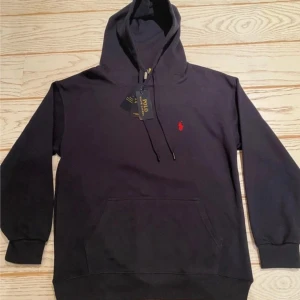 Navy hoodie från Polo Ralph Lauren - Svart hoodie från Polo Ralph Lauren med röd broderad logga på bröstet. Klassisk känguruficka, snörning i huvan och ribbade muddar. Tillverkad i mjuk bomull, perfekt för höst och vinter. Enkel och stilren design som funkar till allt. Qr koden funka 