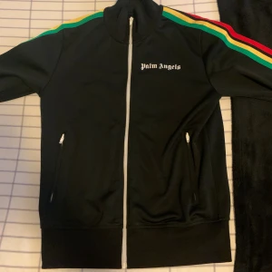 Palm Angels tröja - Svart track jacket från Palm Angels med dragkedja framtill och vita zippar på fickorna. Snygga ränder i grönt, gult och rött längs ärmarna. Hög krage och logga på bröstet. Perfekt för en streetwear-look.