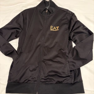Svart EA7 Emporio Armani jacka - Svart jacka och svarta byxor från EA7 Emporio Armani med guldfärgad logga på bröstet. Jackan har hel dragkedja framtill, ståkrage och ribbade muddar vid ärmslut och nederkant. Tillverkad i ett slätt, lätt polyestermaterial som ger en sportig look. Byxor ingår!!